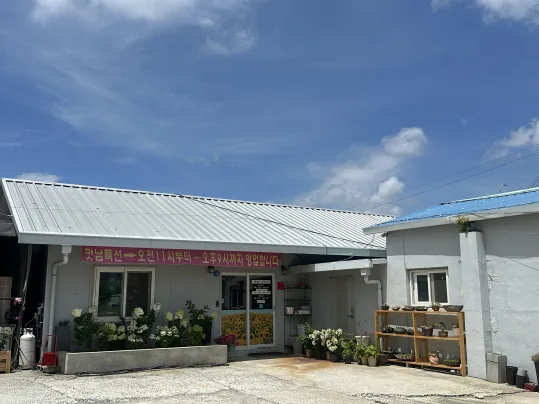 맛남의광장 건물 — 파란 하늘 아래 흰 금속 지붕 단층 건물, 처마 밑 분홍 현수막, 입구 앞 흰 꽃 화분