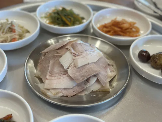 잔치수육 가까이 — 쇠 접시에 겹쳐 담은 얇게 썬 돼지고기 수육, 뒤로 반찬